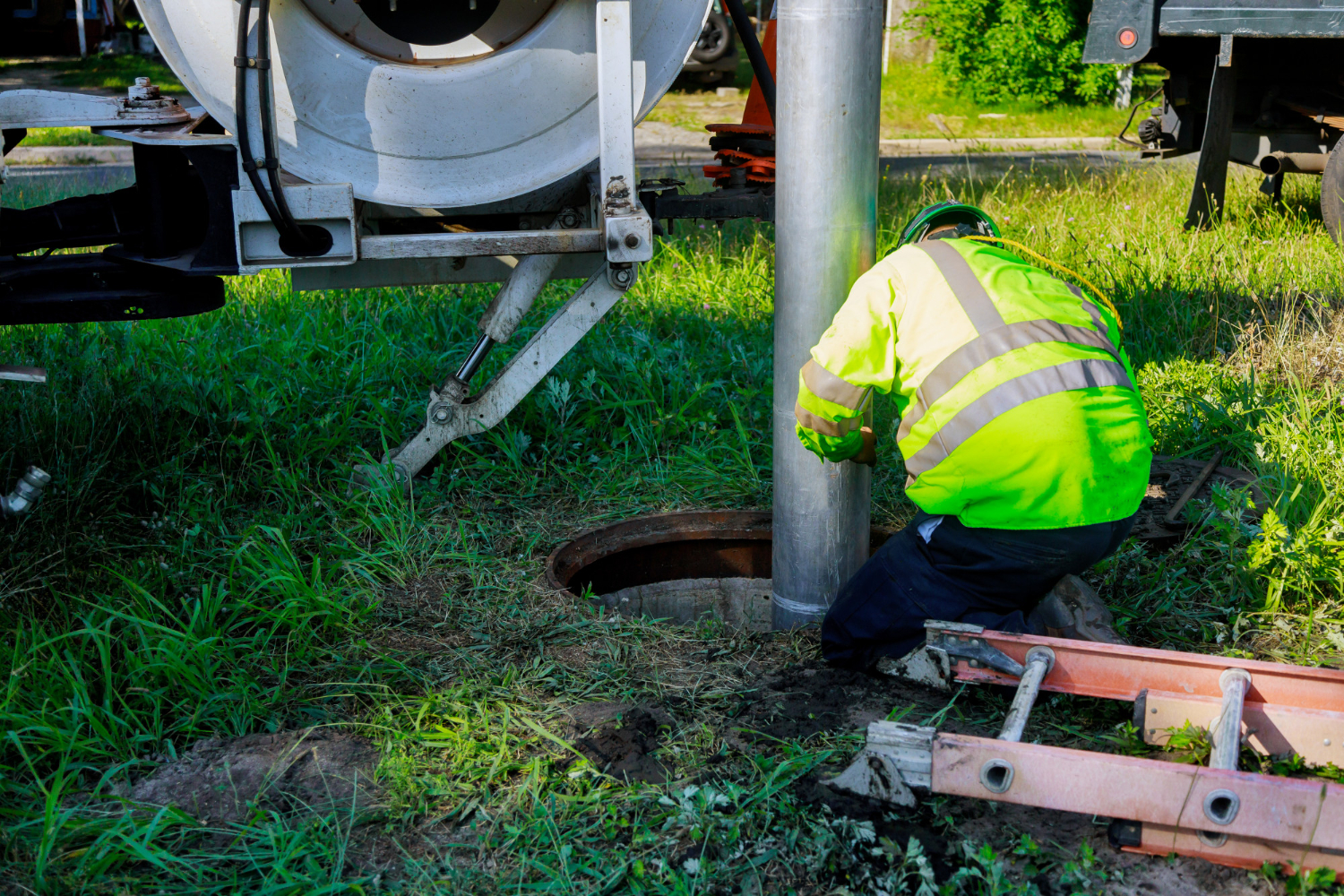 Worker in reflective safety gear performing septic system maintenance, inspecting a septic tank access point in a grassy area, with maintenance equipment nearby.