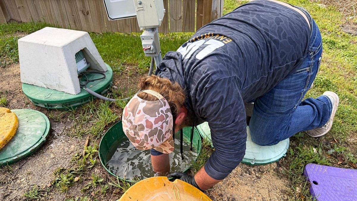 Luna Environmental technician kneeling at an open septic riser, arm deep in the pump tank to pull the pump